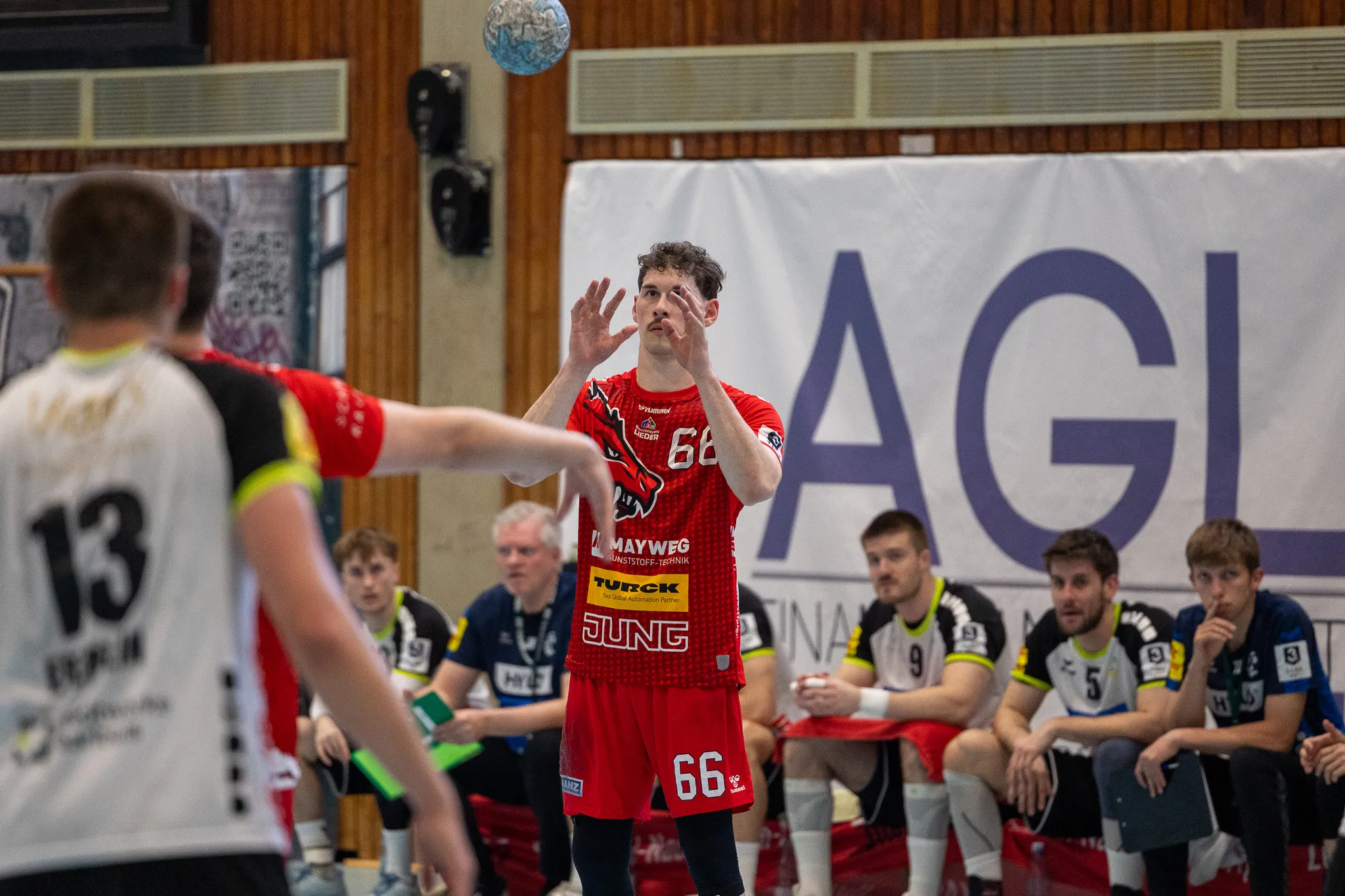 Handballspieler in rotem Trikot wirft den Ball ab, während ein Gegenspieler im Hintergrund versucht, ihn zu blocken. Zuschauer sitzen auf Bänken am Spielfeldrand.