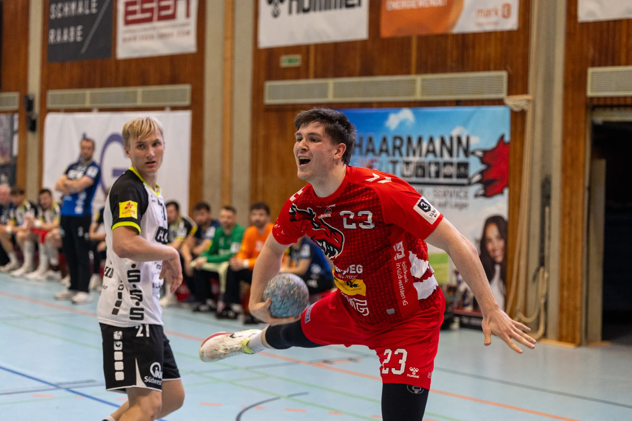 Handballspieler in rotem Trikot mit der Nummer 23 springt jubelnd auf, während ein Gegenspieler im schwarzen Trikot im Hintergrund steht. Im Hintergrund sind weitere Spieler und Zuschauer zu sehen.