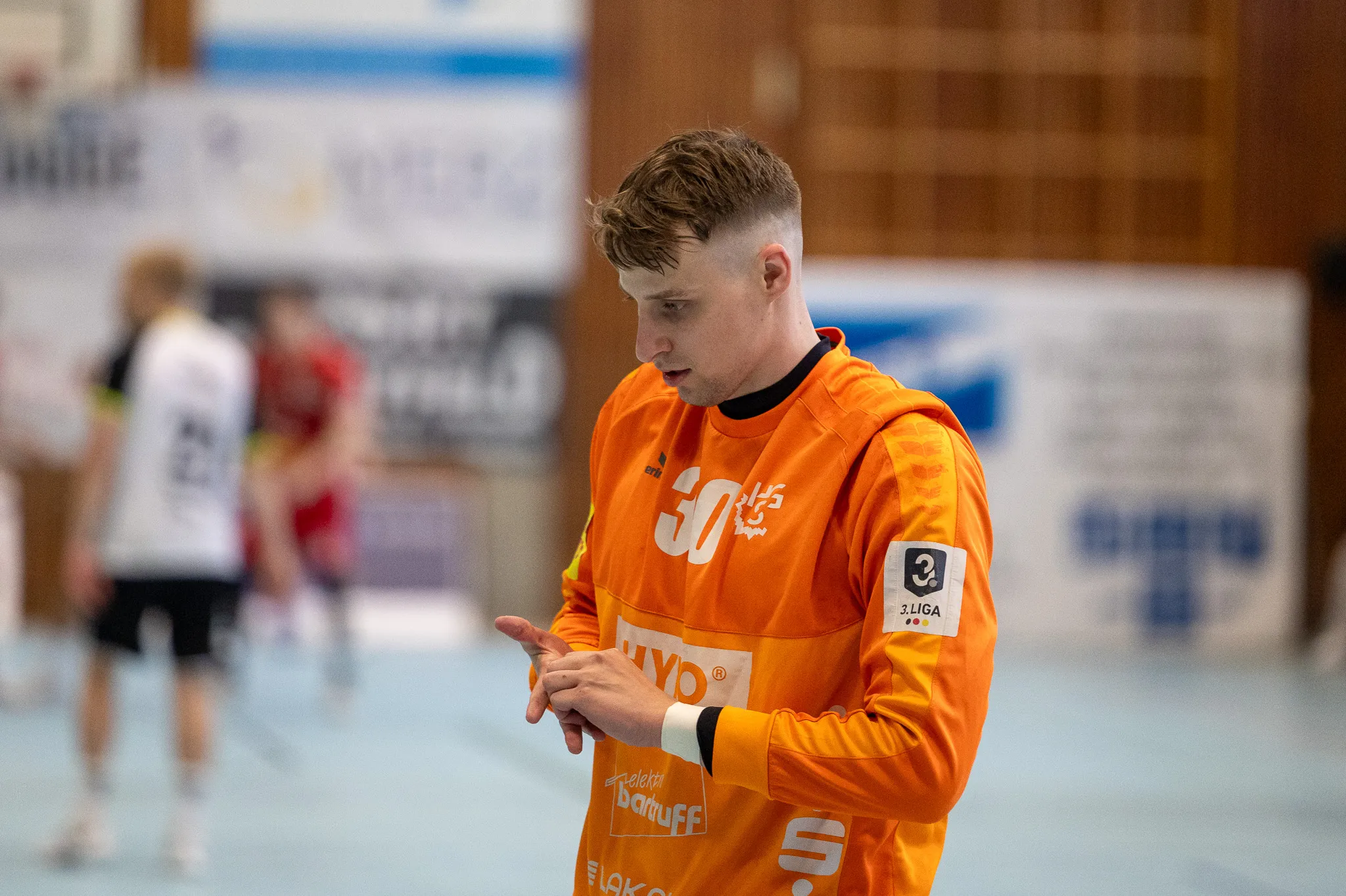 Handballspieler in orangefarbenem Trikot mit der Nummer 30 konzentriert sich auf seine Hände, während im Hintergrund ein Spiel stattfindet.