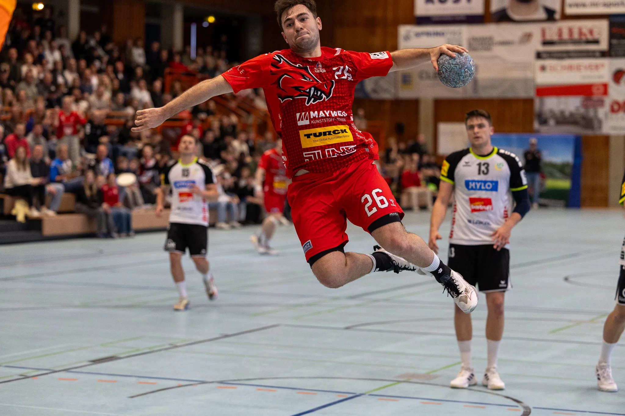 Handballspieler in rotem Trikot springt beim Wurf, während ein Gegenspieler im Hintergrund steht. Im Hintergrund sind Zuschauer auf den Rängen zu sehen.