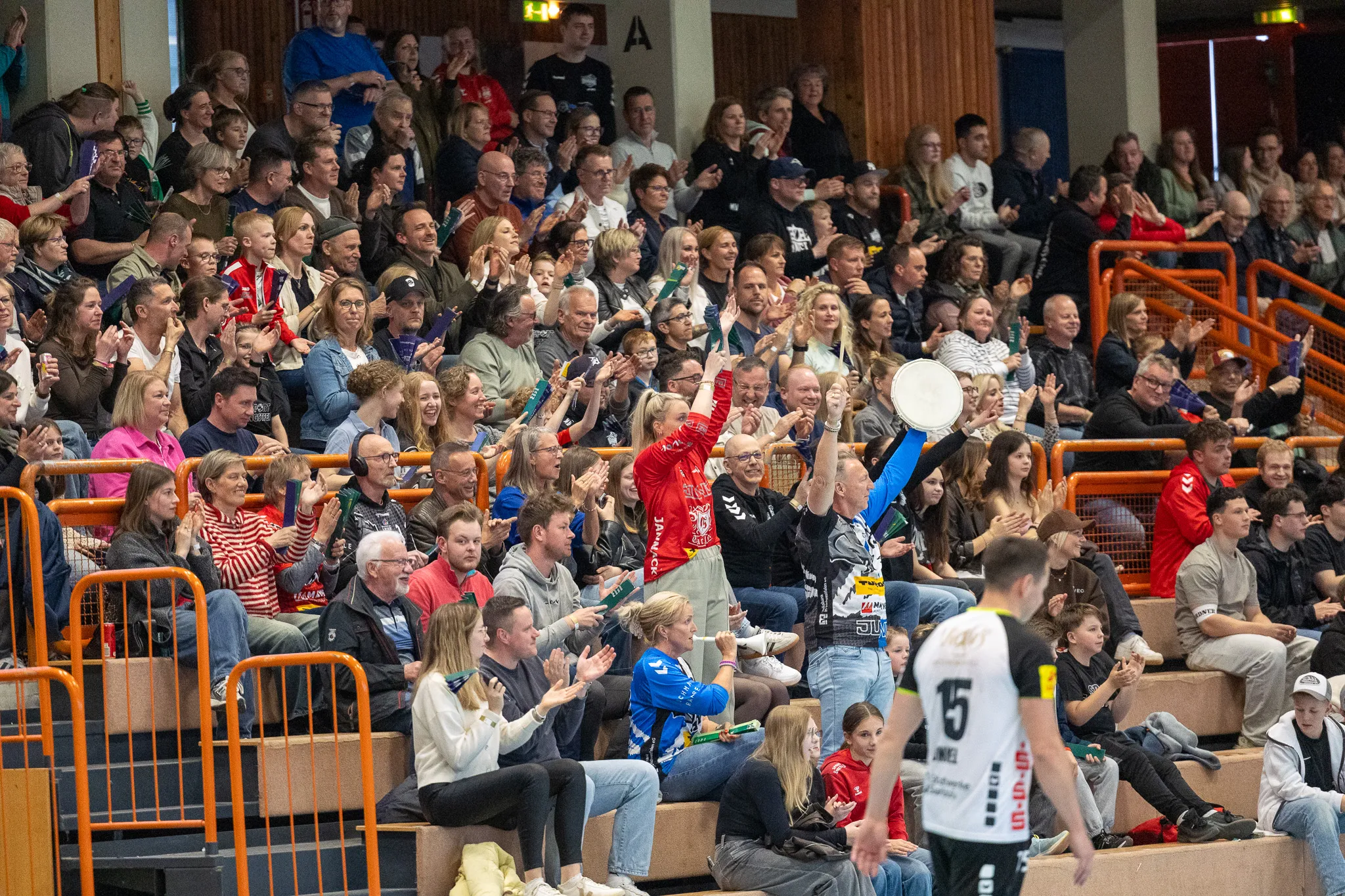 Zuschauer jubeln und klatschen bei einem Handballspiel, einige halten Fanartikel hoch. Im Vordergrund ein Spieler mit der Trikotnummer 15.