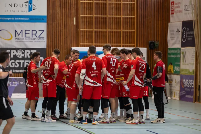 Handballspieler der SGSH Dragons stehen in roter Spielkleidung im Kreis und besprechen Taktiken, während sie Wasserflaschen halten.