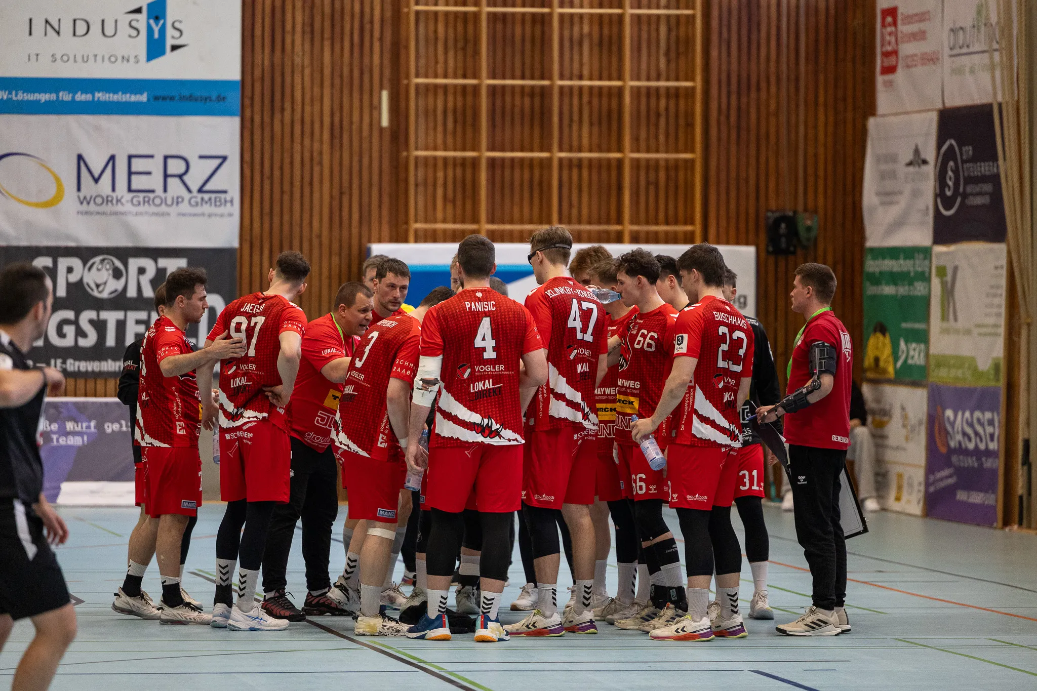 Handballspieler der SGSH Dragons stehen in roter Spielkleidung im Kreis und besprechen Taktiken, während sie Wasserflaschen halten.
