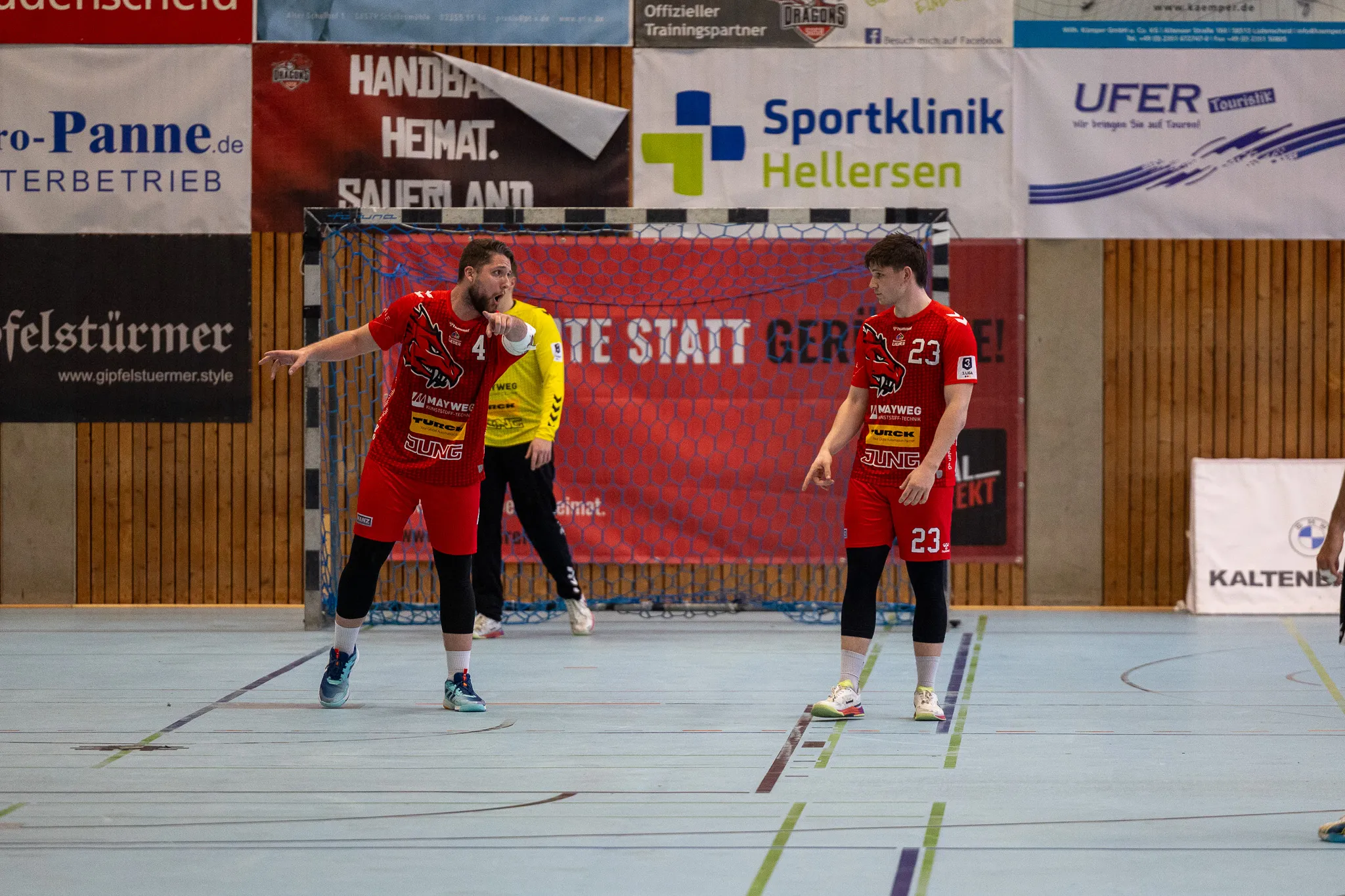 Zwei Handballspieler in roten und schwarzen Trikots stehen auf einem Hallenparkett, der Spieler links deutet mit ausgestrecktem Arm nach vorne.