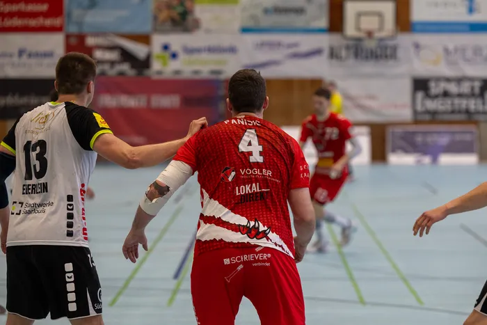 Zwei Handballspieler kämpfen um den Ball: Ein Spieler in rotem Trikot mit der Nummer 4 und ein Spieler in schwarzem Trikot mit der Nummer 13.