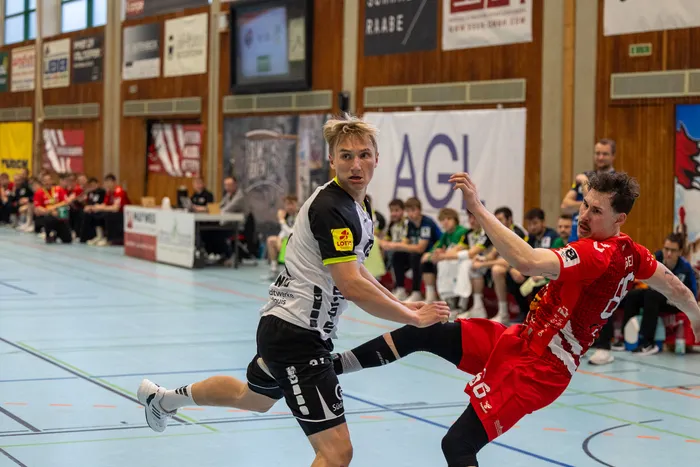 Zwei Handballspieler im Spiel: Ein Spieler in Schwarz bereitet einen Wurf vor, während ein Spieler in Rot versucht, den Wurf zu blocken. Im Hintergrund sind weitere Spieler und Zuschauer in einer Sporthalle zu sehen.