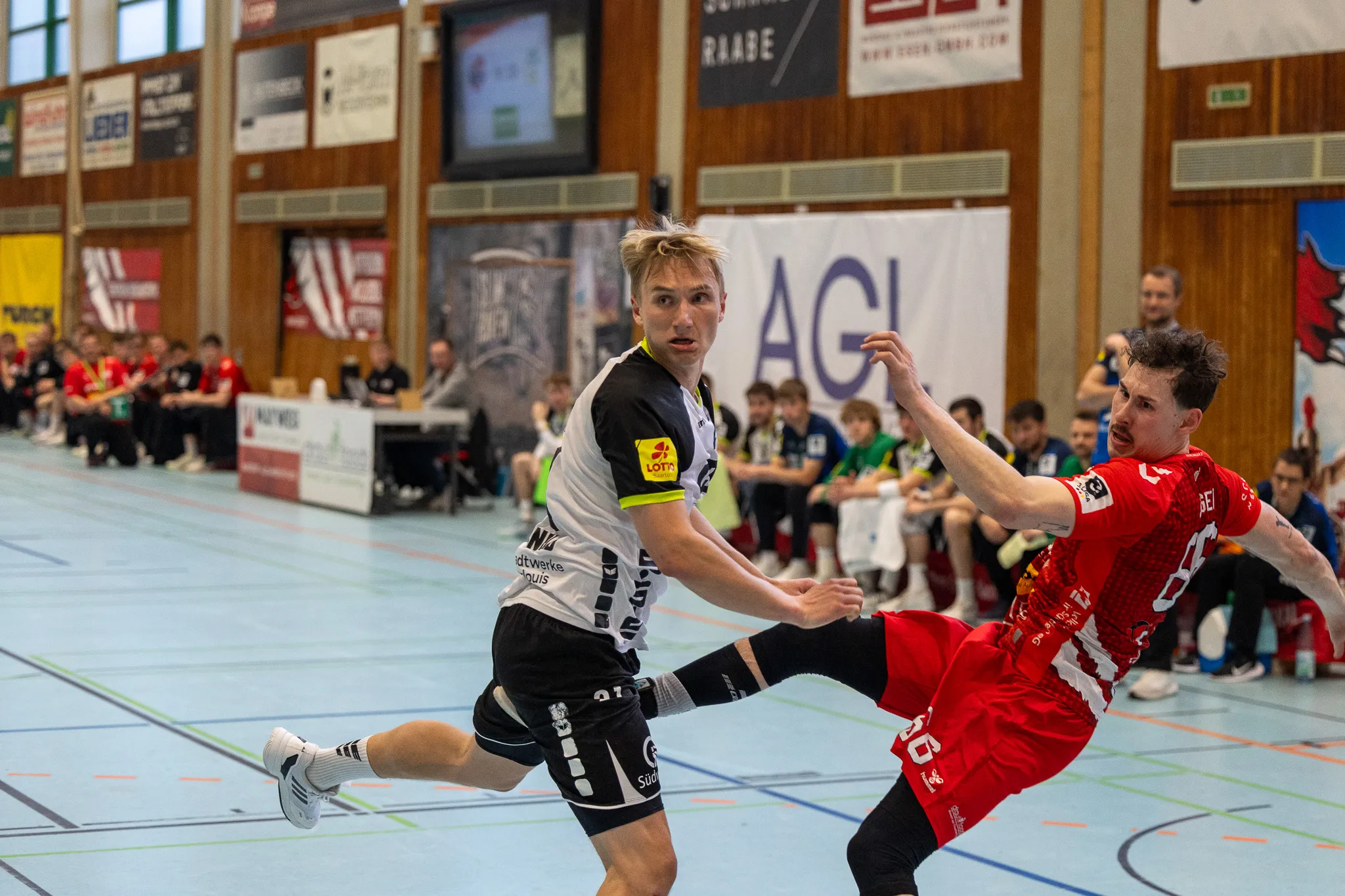 Zwei Handballspieler im Spiel: Ein Spieler in Schwarz bereitet einen Wurf vor, während ein Spieler in Rot versucht, den Wurf zu blocken. Im Hintergrund sind weitere Spieler und Zuschauer in einer Sporthalle zu sehen.