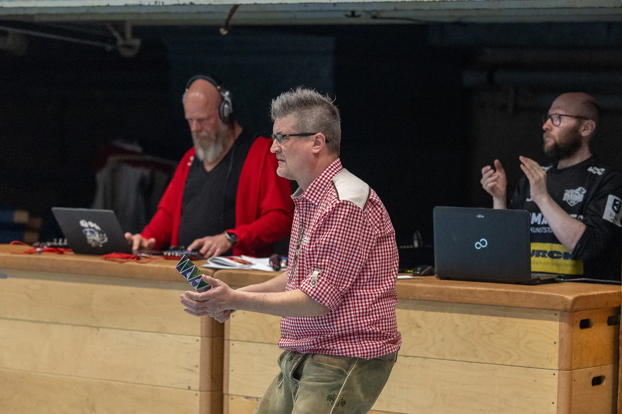 Mann in rot-kariertem Hemd hält Spielkarten vor sich, während zwei weitere Männer im Hintergrund an Laptops arbeiten.