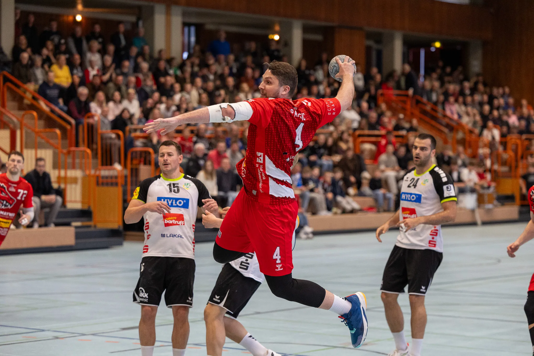 Handballspieler in roten Trikots springt zum Wurf, während Gegenspieler in schwarzen und blauen Trikots ihn verteidigen. Im Hintergrund eine gefüllte Tribüne.