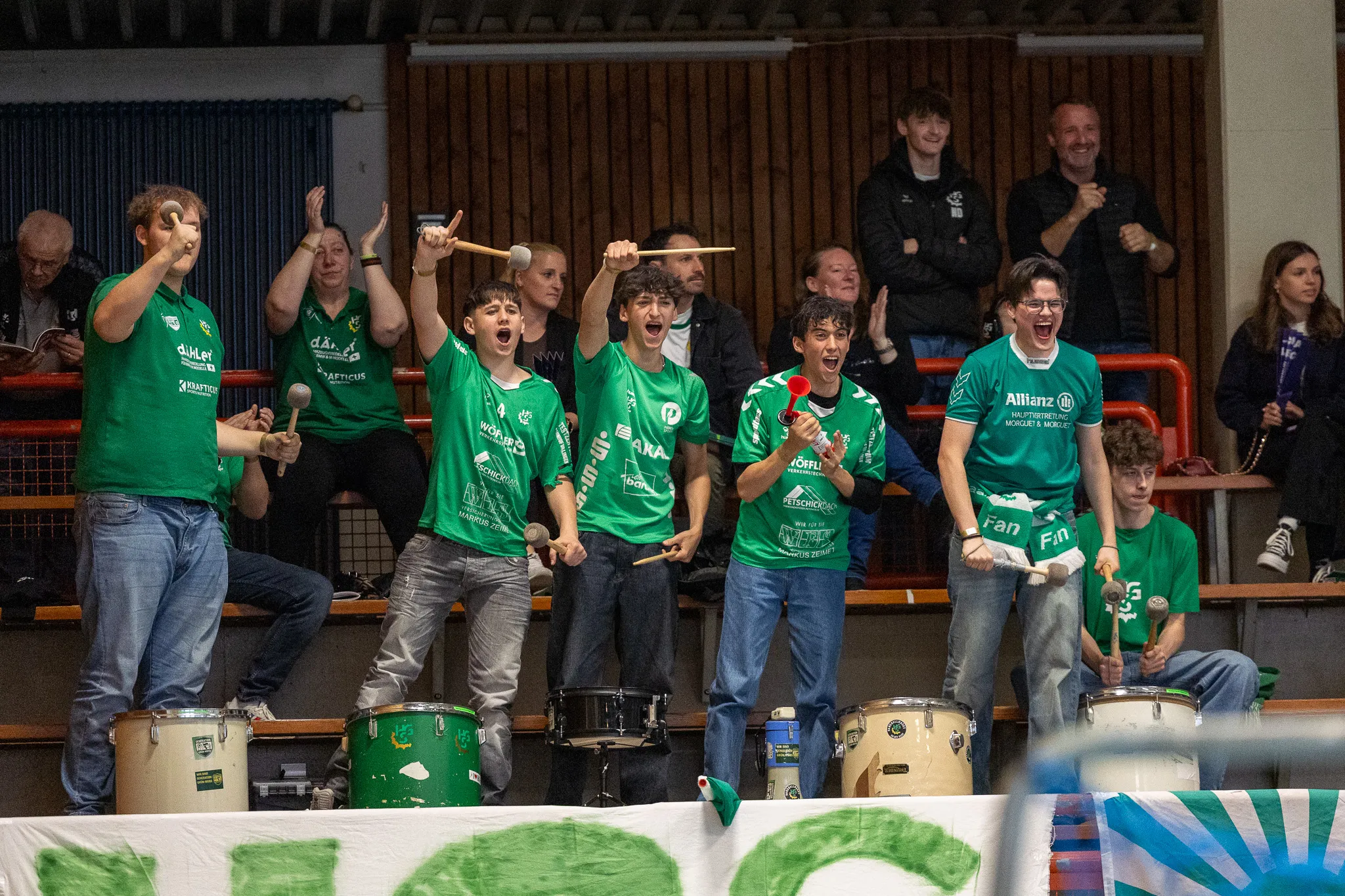 Jubelnde Fans in grünen T-Shirts schlagen auf Trommeln und heben Arme in die Luft, während im Hintergrund weitere Zuschauer zu sehen sind.