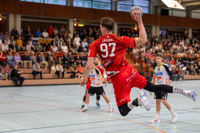 Handballspieler in rotem Trikot mit der Nummer 92 springt zum Wurf, während ein Gegenspieler in blauem Trikot ihn blockt. Im Hintergrund sind Zuschauer in einer Sporthalle zu sehen.