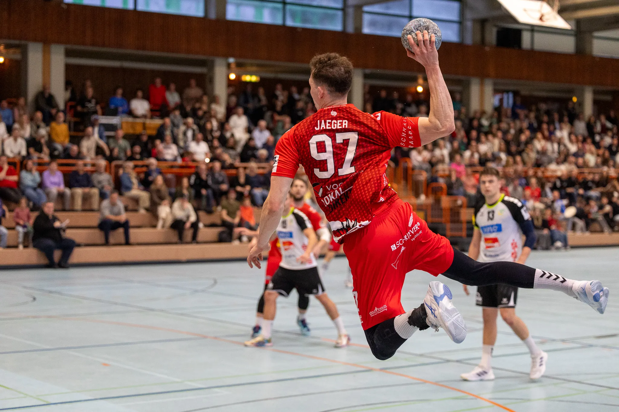 Handballspieler in rotem Trikot mit der Nummer 92 springt zum Wurf, während ein Gegenspieler in blauem Trikot ihn blockt. Im Hintergrund sind Zuschauer in einer Sporthalle zu sehen.