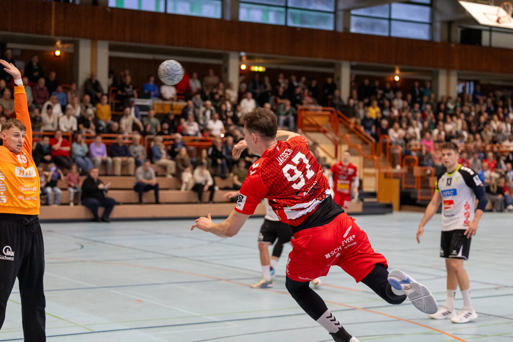 Handballspieler in rotem Trikot mit der Nummer 92 springt zum Wurf, während ein Gegenspieler ihn verteidigt. Im Hintergrund sind Zuschauer in einer Sporthalle zu sehen.