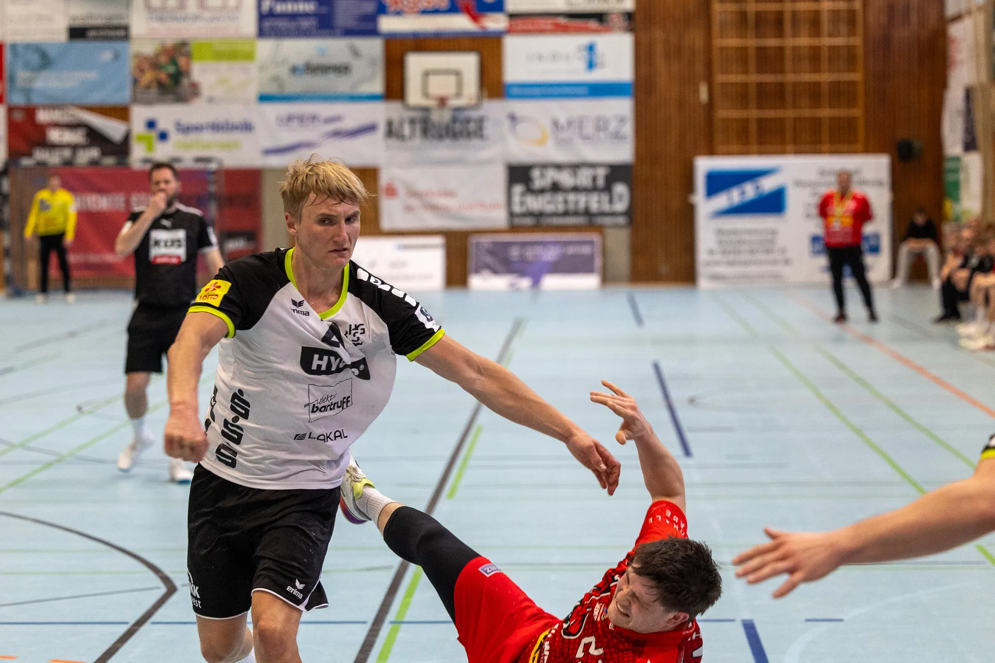 Handballspieler in schwarzem Trikot mit Werbeaufdrucken versucht, einen Pass über einen Gegner im roten Trikot zu spielen. Im Hintergrund sind Zuschauer und Werbebanner in einer Sporthalle zu sehen.