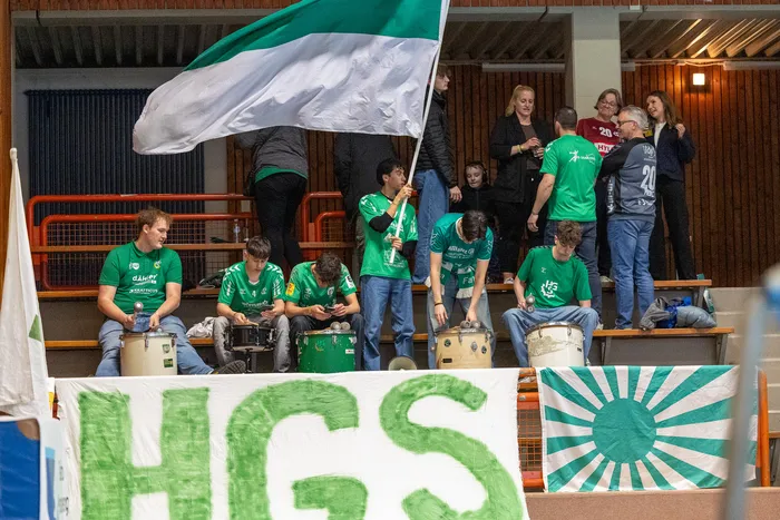 Mehrere Trommler in grünen T-Shirts spielen vor Zuschauern in einer Sporthalle. Im Hintergrund weht eine schwarz-weiß-grüne Flagge.