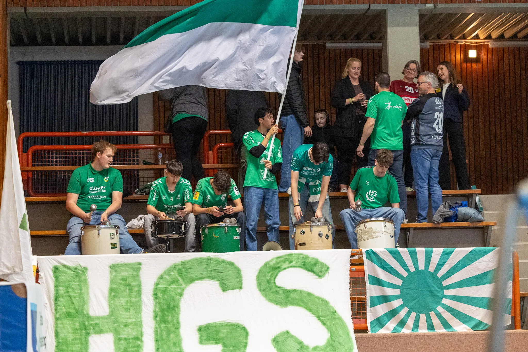Mehrere Trommler in grünen T-Shirts spielen vor Zuschauern in einer Sporthalle. Im Hintergrund weht eine schwarz-weiß-grüne Flagge.