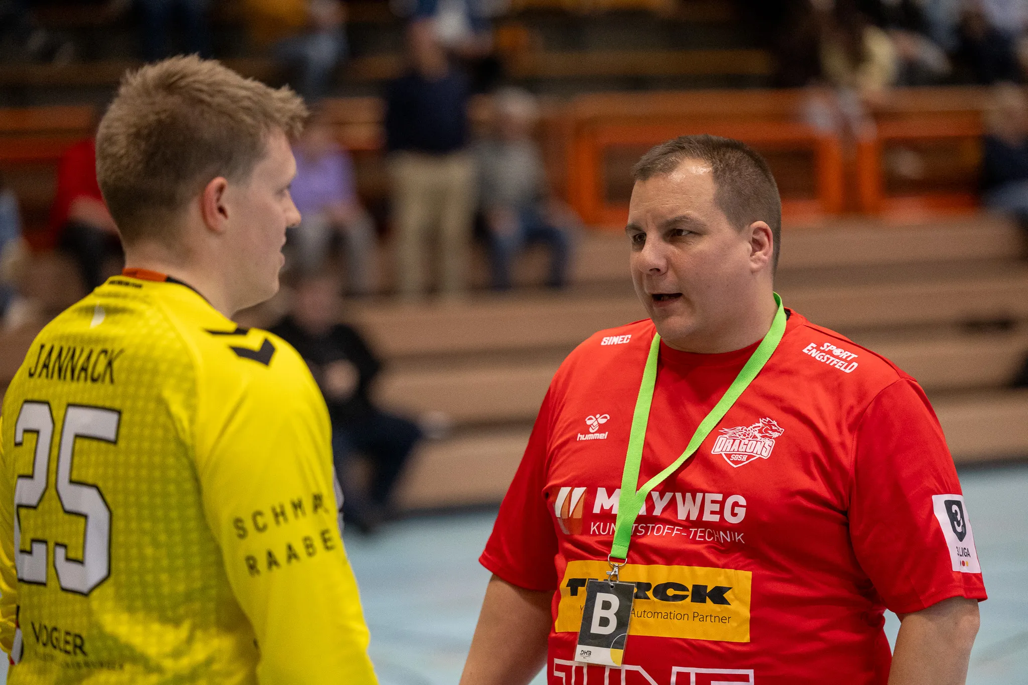 Zwei Handballspieler stehen sich auf dem Spielfeld gegenüber. Der Spieler im roten Trikot trägt eine Medaille um den Hals.