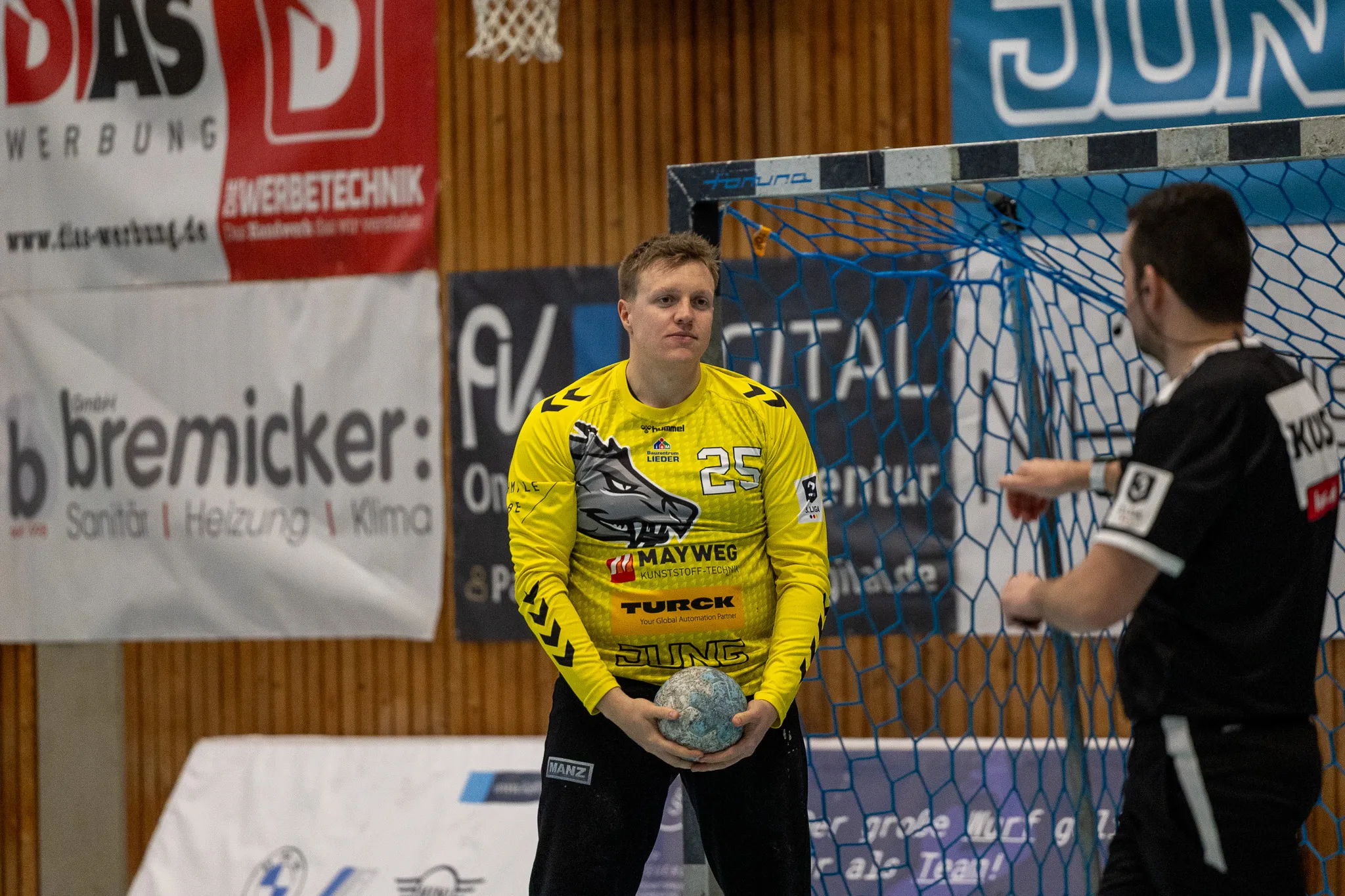 Handballtorwart in gelbem Trikot mit der Nummer 25 hält den Ball vor sich, während ein Gegenspieler im Hintergrund zu sehen ist.