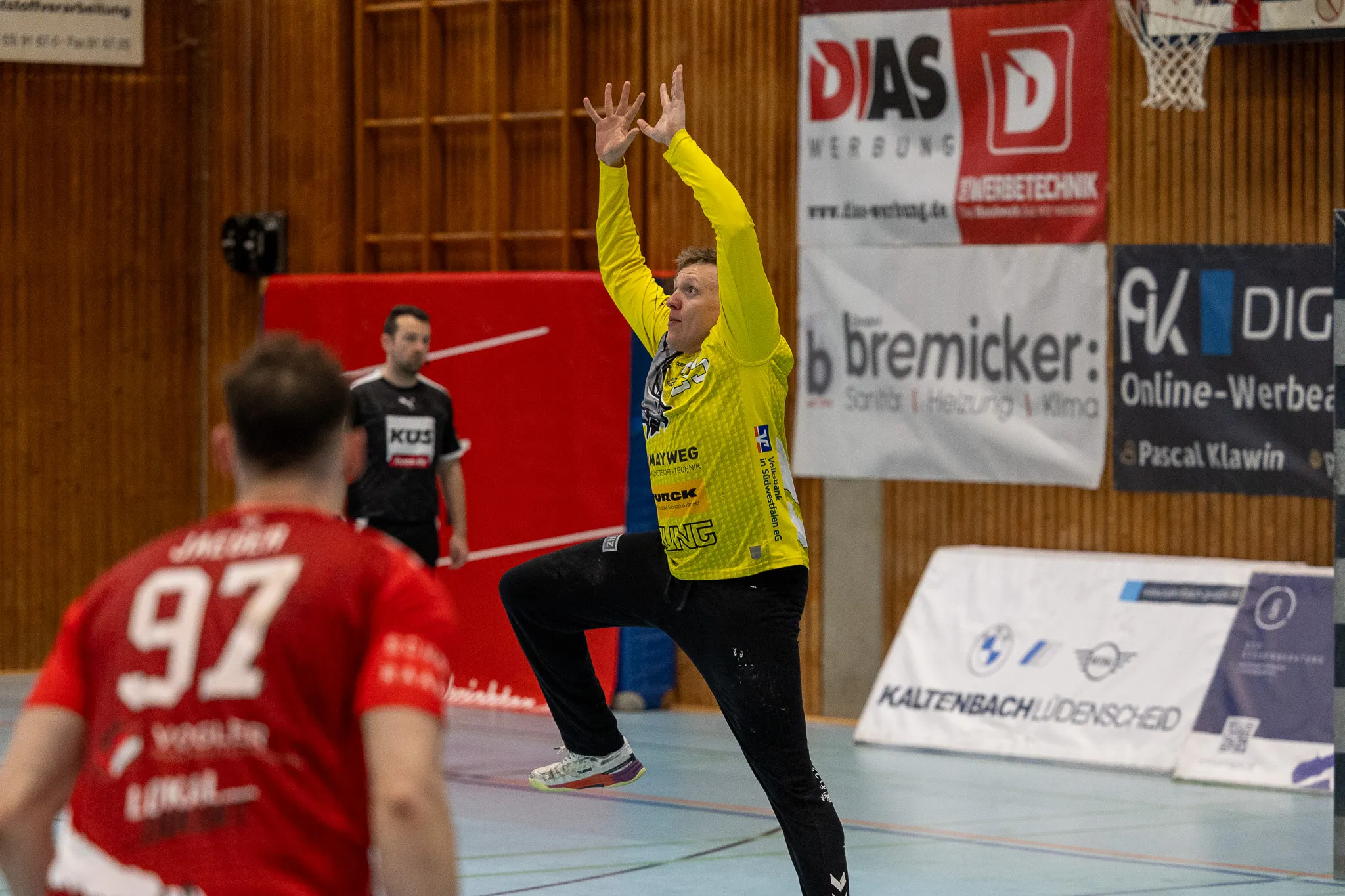 Handballspieler in gelbem Trikot wirft den Ball während eines Spiels, ein weiterer Spieler in rotem Trikot bereitet sich auf die Verteidigung vor.