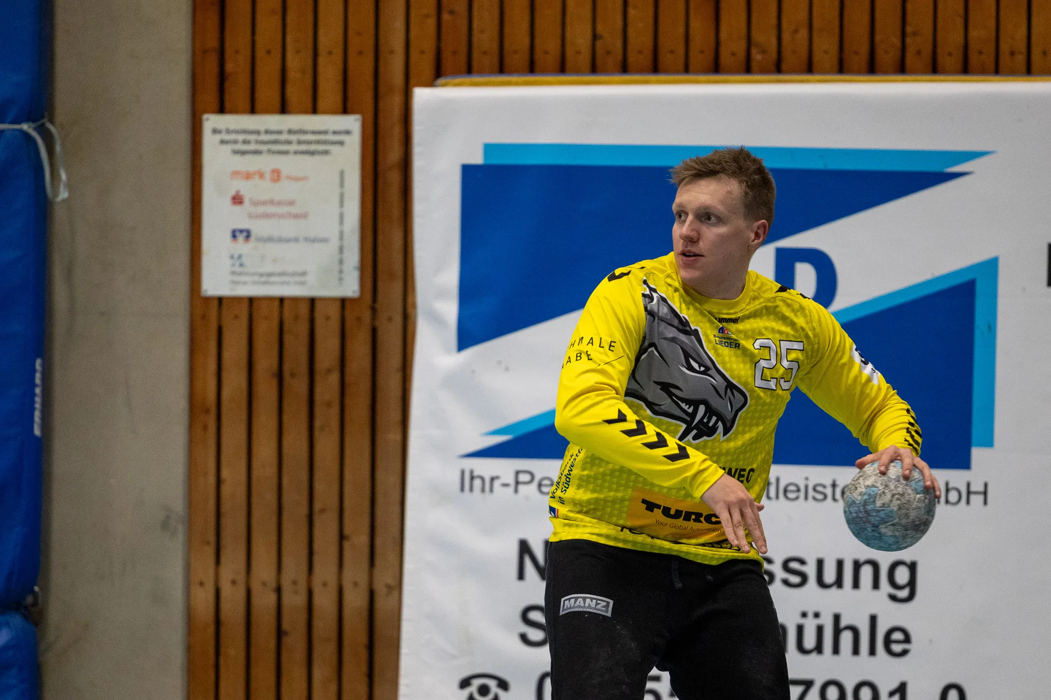 Handballspieler in gelbem Trikot mit der Nummer 25 hält Ball in der Hand während des Spiels in einer Sporthalle.