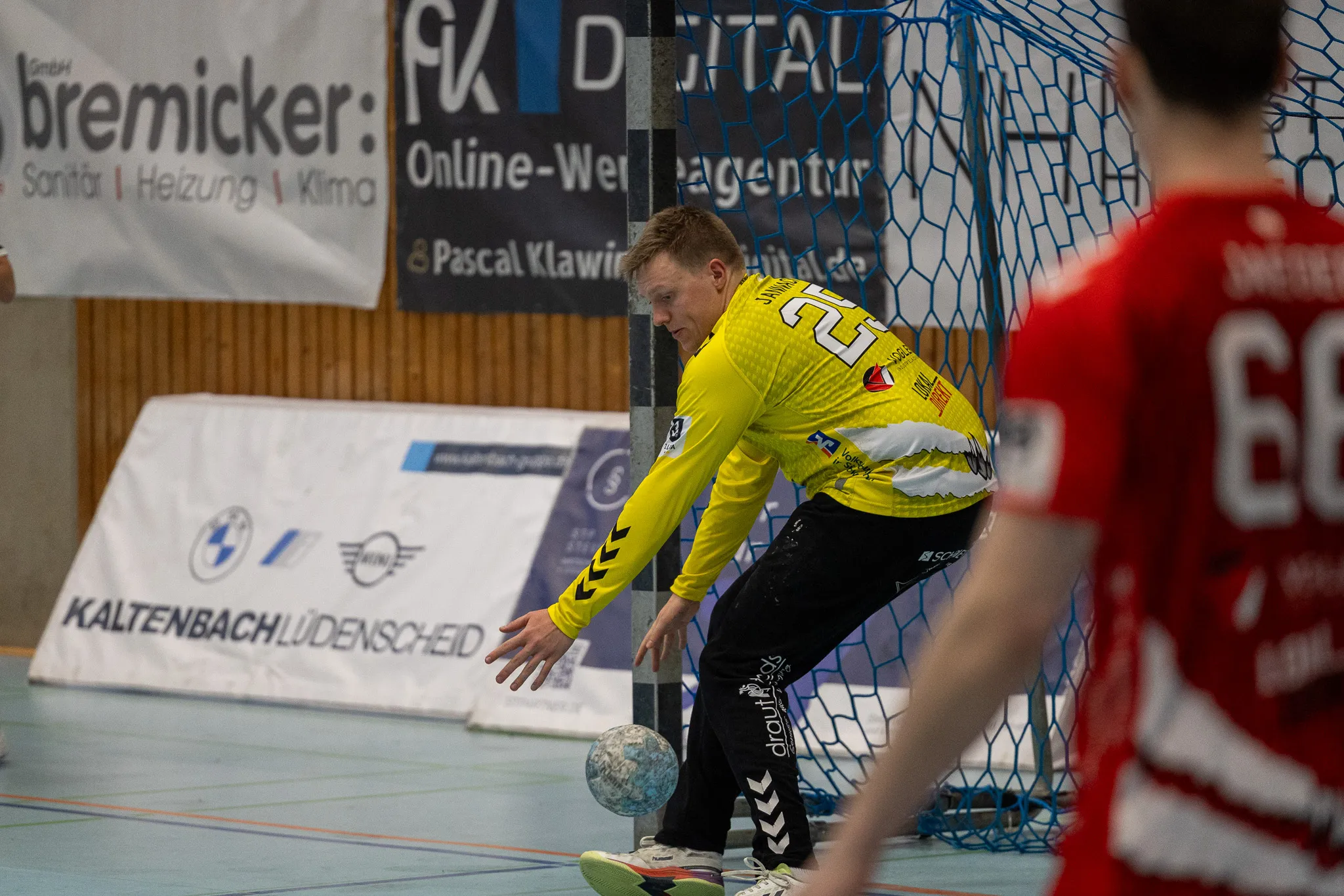 Handballspieler in gelbem Trikot bereitet einen Wurf vor, während ein Gegenspieler im roten Trikot im Hintergrund steht. Im Hintergrund sind Werbebanner für lokale Unternehmen zu sehen.