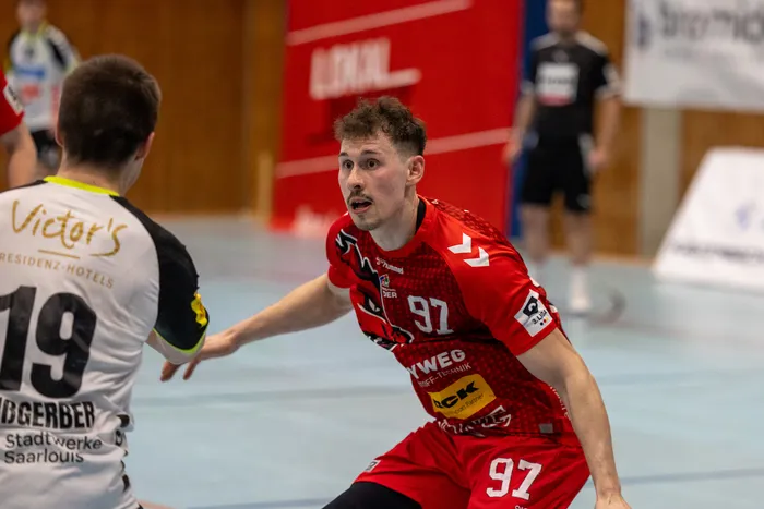Handballspieler mit der Trikotnummer 97 in Rot und Weiß bereitet einen Wurf vor, während ein Gegenspieler im Hintergrund zu sehen ist.