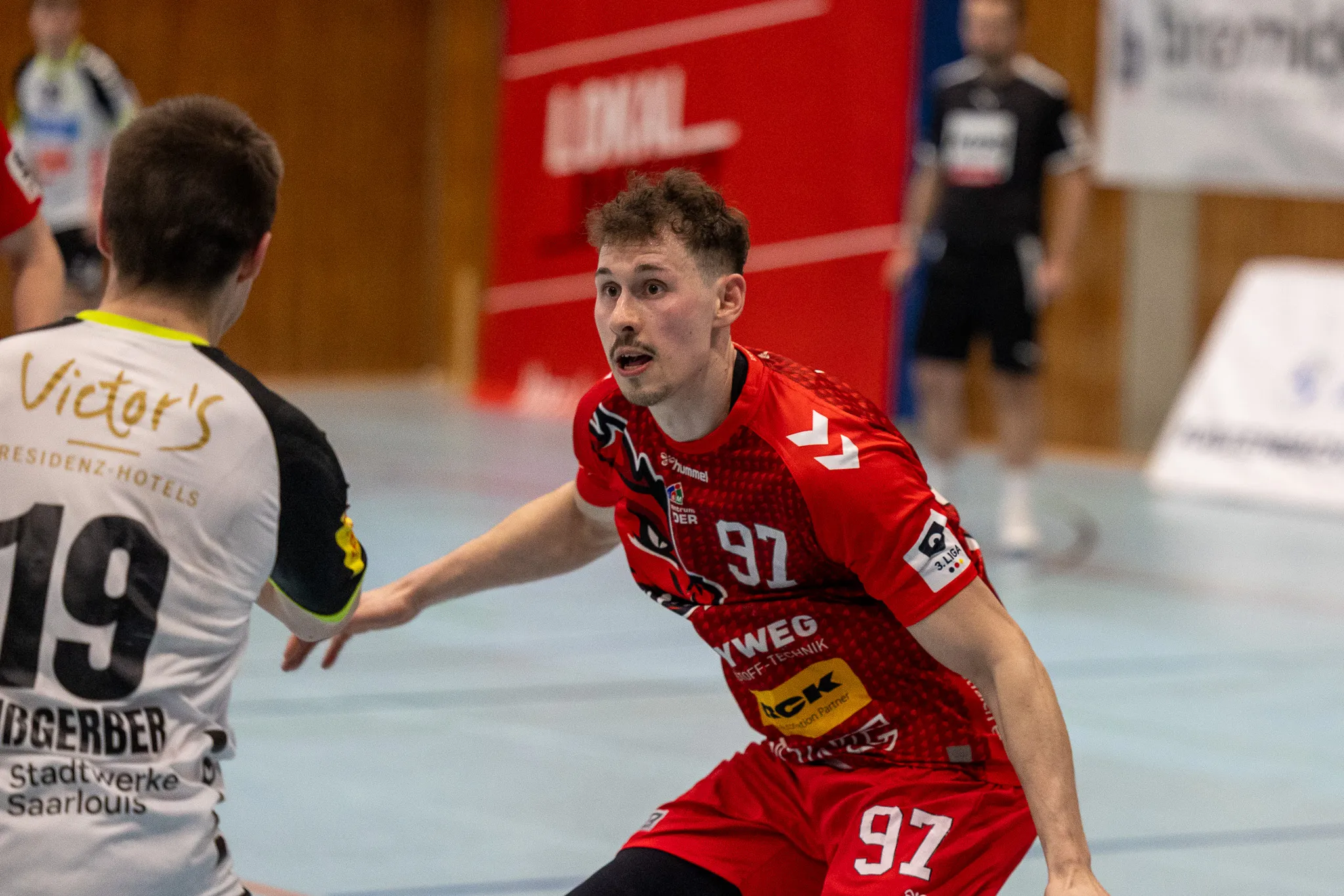 Handballspieler mit der Trikotnummer 97 in Rot und Weiß bereitet einen Wurf vor, während ein Gegenspieler im Hintergrund zu sehen ist.
