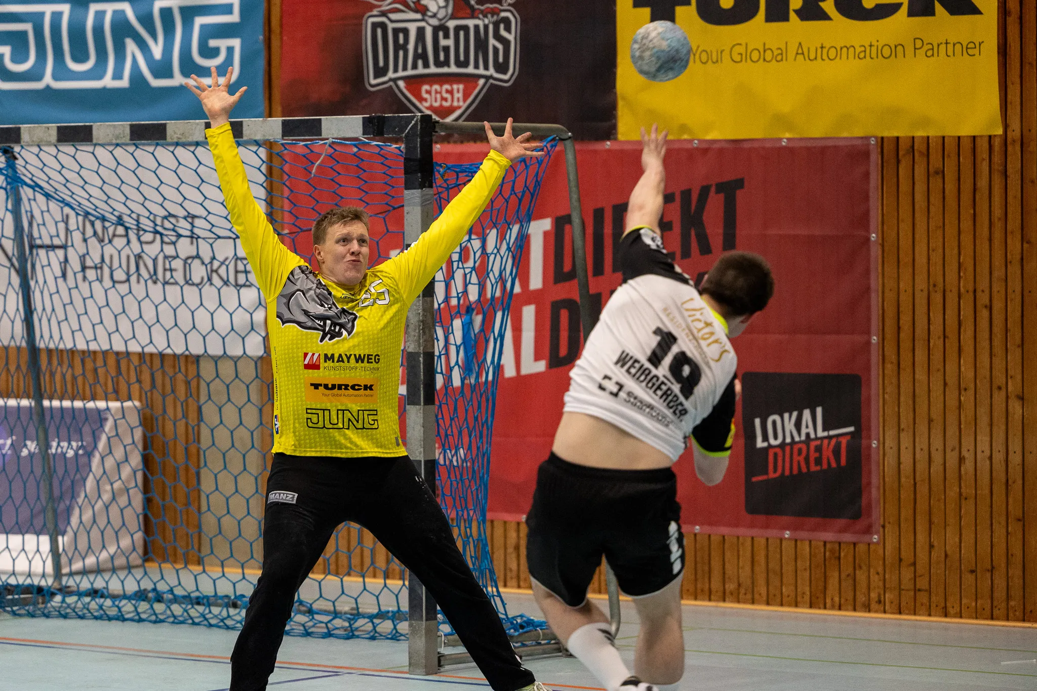 Torwart in gelber Ausrüstung streckt die Arme aus, um einen Ball zu blocken, während ein gegnerischer Spieler im Hintergrund zum Wurf ausholt.