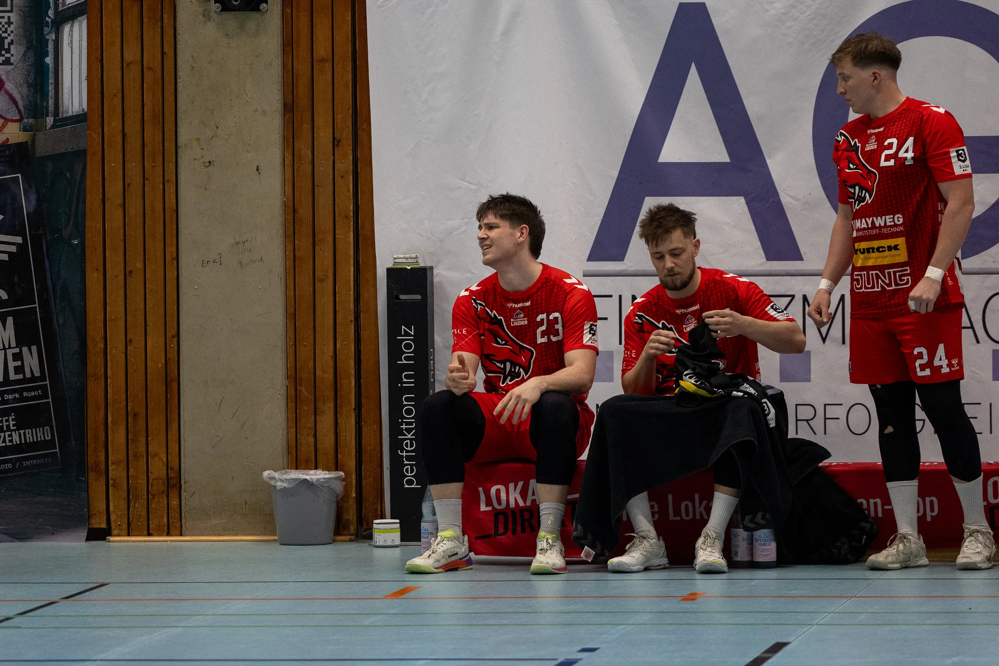Drei Handballspieler der SGSH Dragons sitzen auf einer Bank in der Sporthalle, zwei blicken nach vorn, einer nach unten. Sie tragen rote Trikots mit der Nummer 23 und 24.