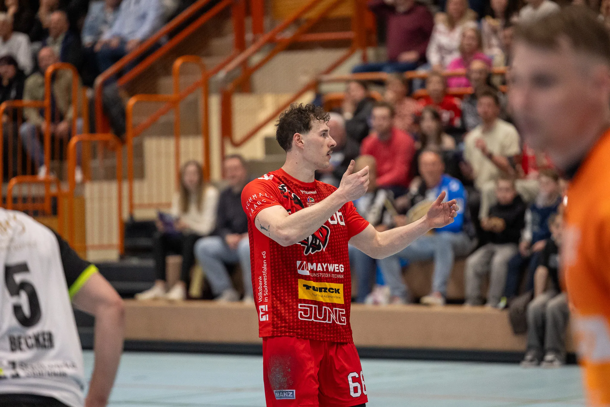 Handballspieler in rotem Trikot mit der Nummer 66 gestikuliert während des Spiels auf dem Spielfeld, im Hintergrund Zuschauer auf den Tribünen.