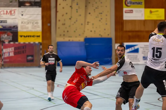 Zwei Handballspieler kämpfen um den Ball, während ein dritter im Hintergrund zu sehen ist. Der Spieler im roten Trikot wird vom Spieler im blauen Trikot attackiert.