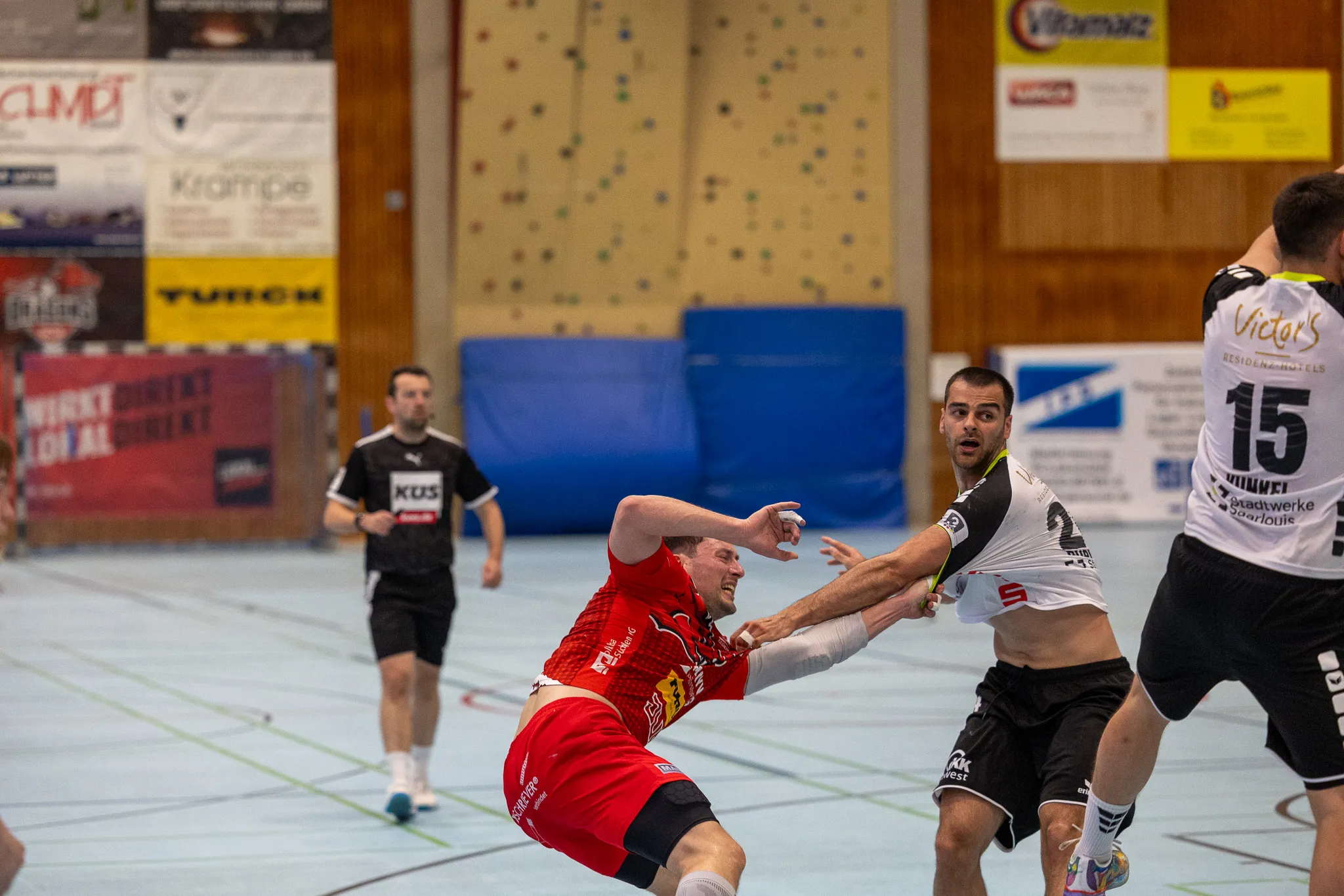 Zwei Handballspieler kämpfen um den Ball, während ein dritter im Hintergrund zu sehen ist. Der Spieler im roten Trikot wird vom Spieler im blauen Trikot attackiert.