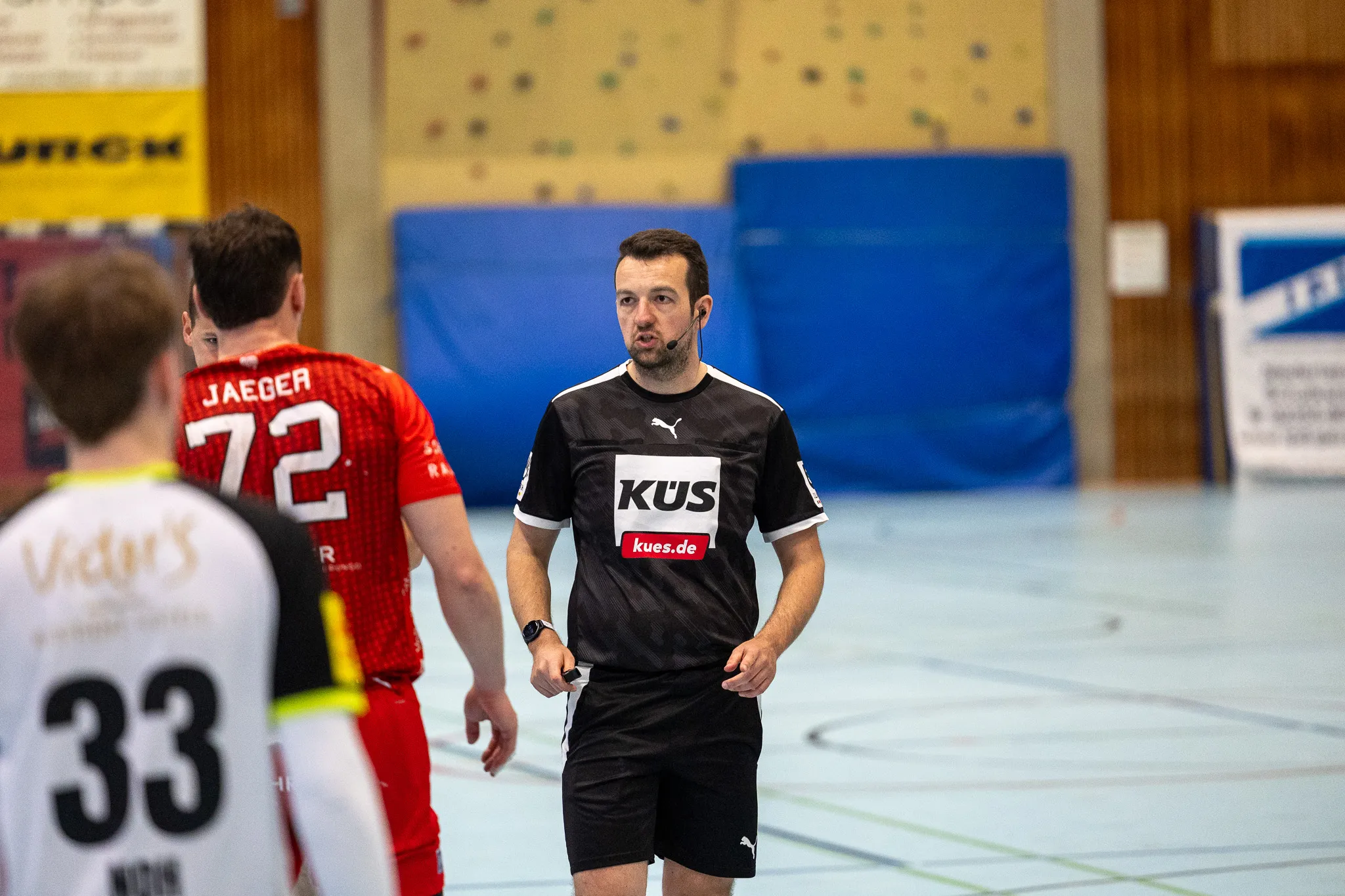 Schiedsrichter mit Headset steht auf einem Handballfeld zwischen zwei Spielern, einer trägt Trikot mit der Nummer 72 und der andere mit der Nummer 33.
