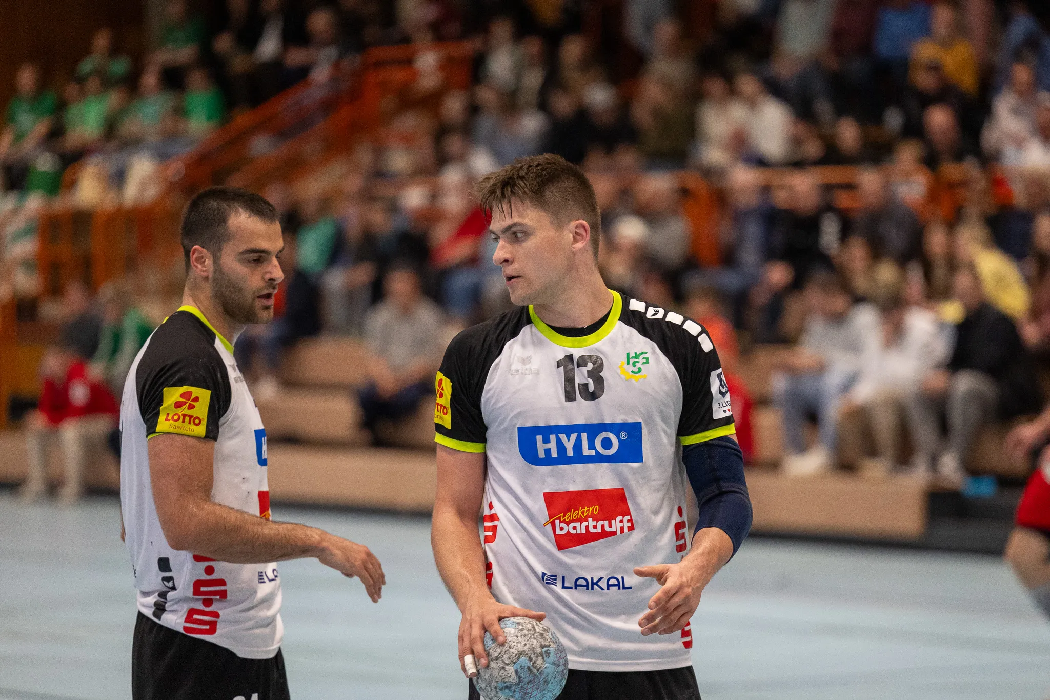 Zwei Handballspieler im Spiel, einer dribbelt den Ball, der andere bereitet sich auf die Verteidigung vor. Im Hintergrund sind Zuschauer auf den Rängen zu sehen.