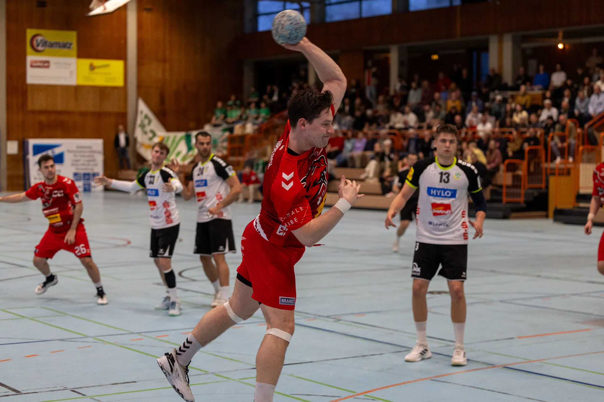 Handballspieler in roten Trikots wirft den Ball während eines Spiels in einer Sporthalle, im Hintergrund Zuschauer.
