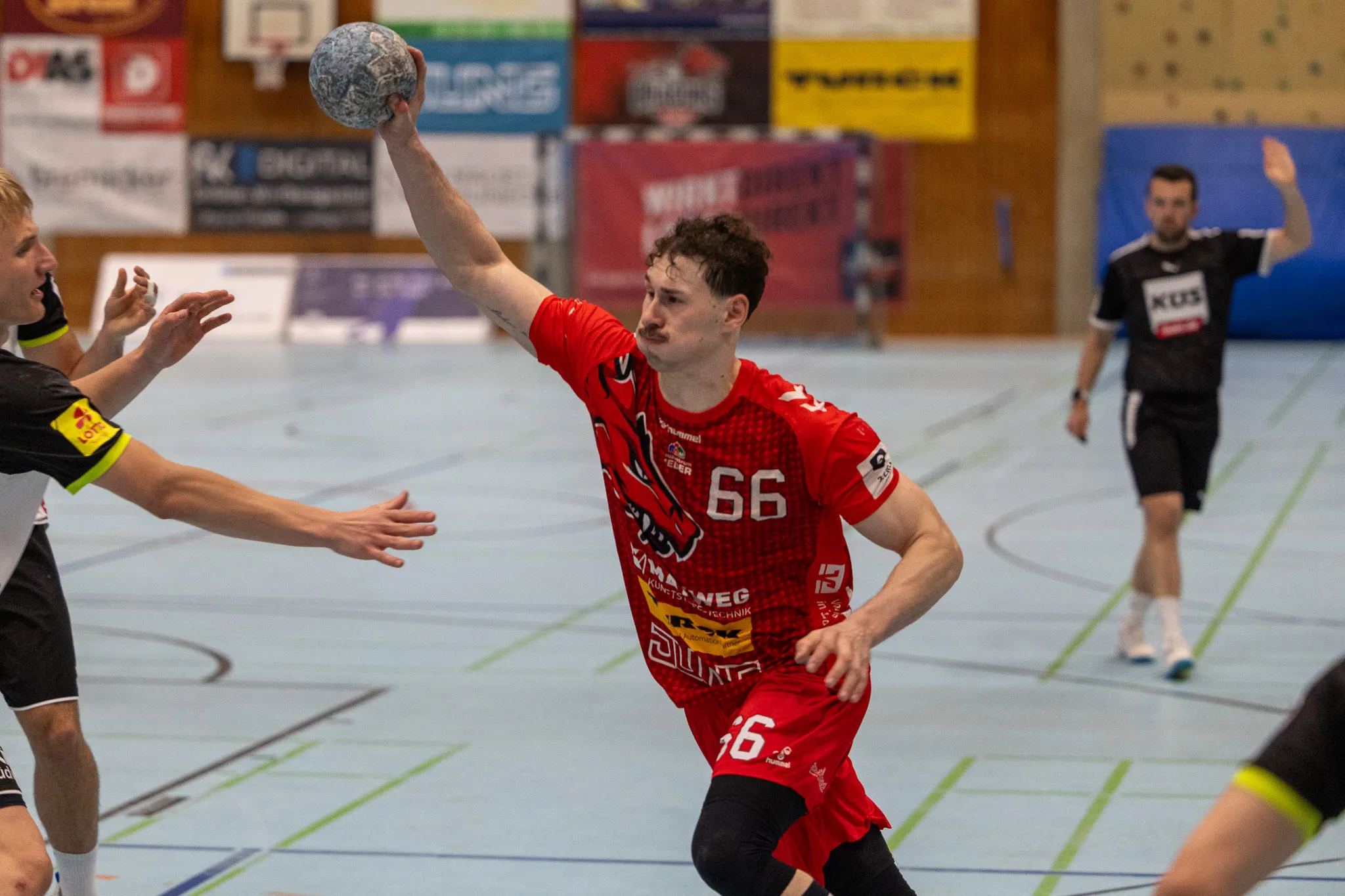 Handballspieler in rotem Trikot mit der Nummer 66 wirft den Ball während eines Spiels. Im Hintergrund sind weitere Spieler und Zuschauer zu sehen.