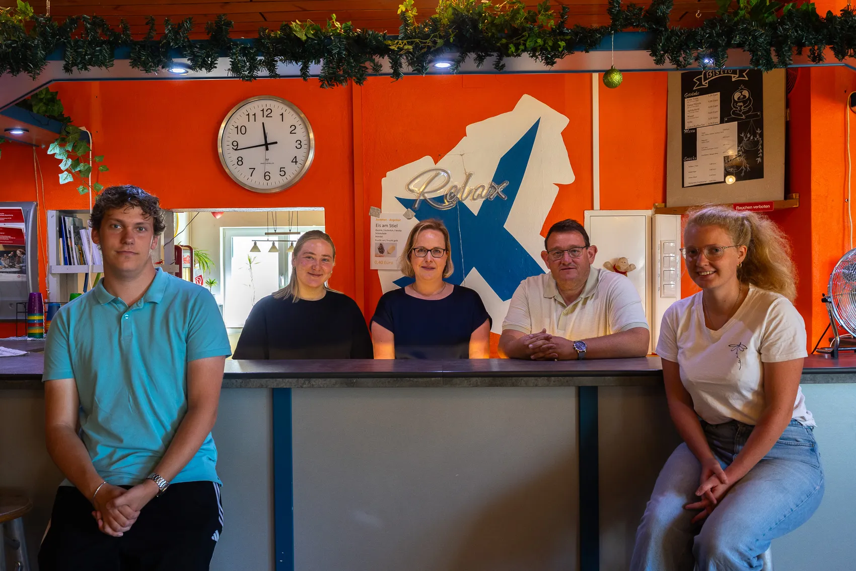 Das Team des Jugendzentrums Kierspe posiert hinter einem Tresen vor einer orangefarbenen Wand mit einer Wanduhr und einem „Relax“-Schriftzug.