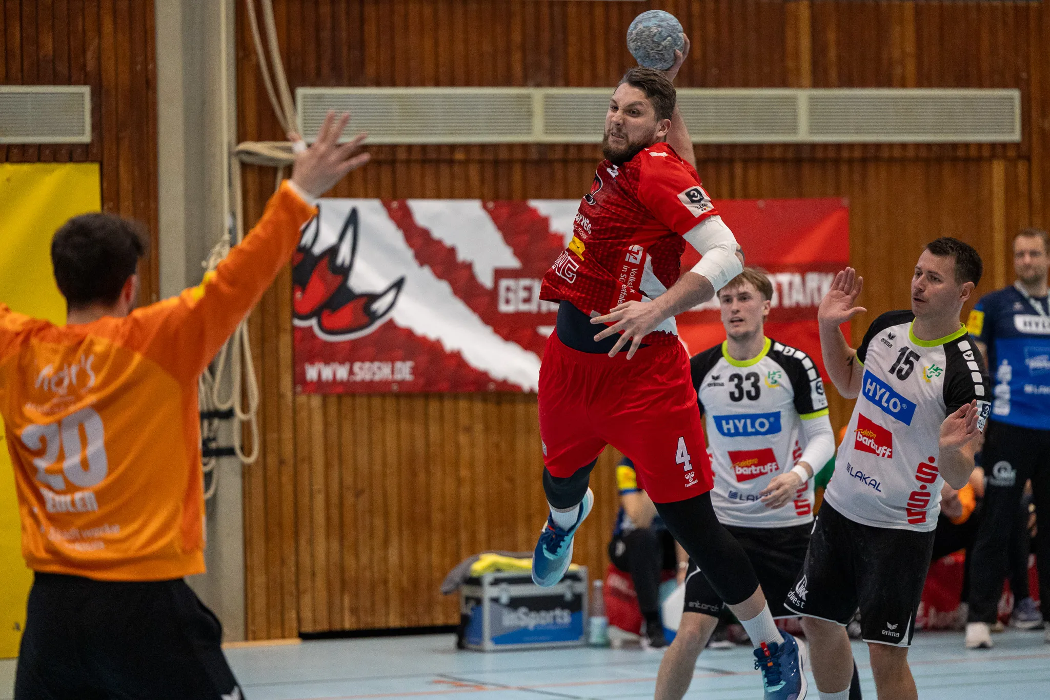 Handballspieler in roten Trikots springen während eines Spiels in der Halle um den Ball. Im Hintergrund ein Werbebanner und weitere Spieler.