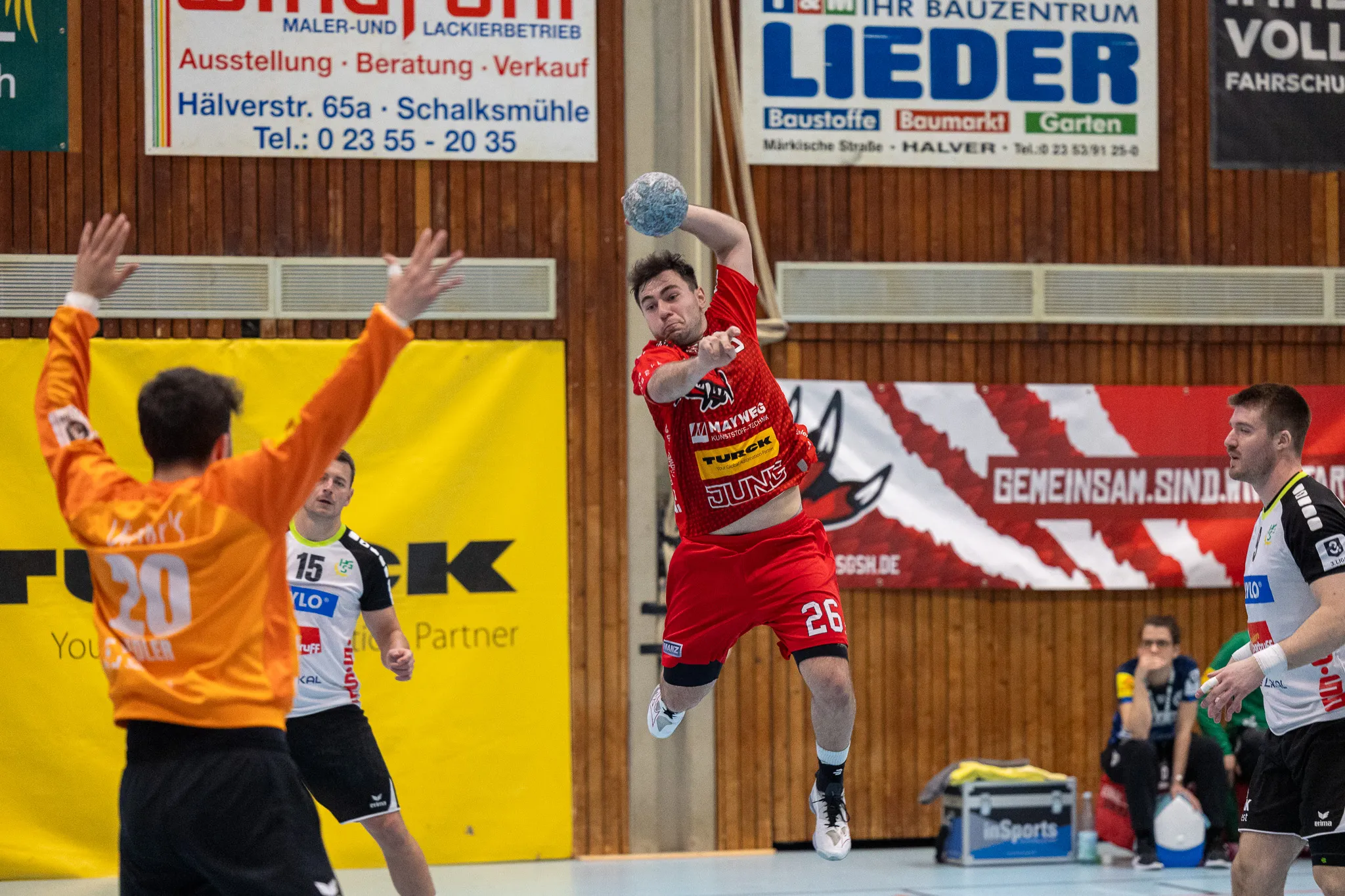 Handballspieler in Rot springt zum Wurf, während Gegenspieler in Orange versuchen, ihn zu blocken. Im Hintergrund Werbebanner und Zuschauer.