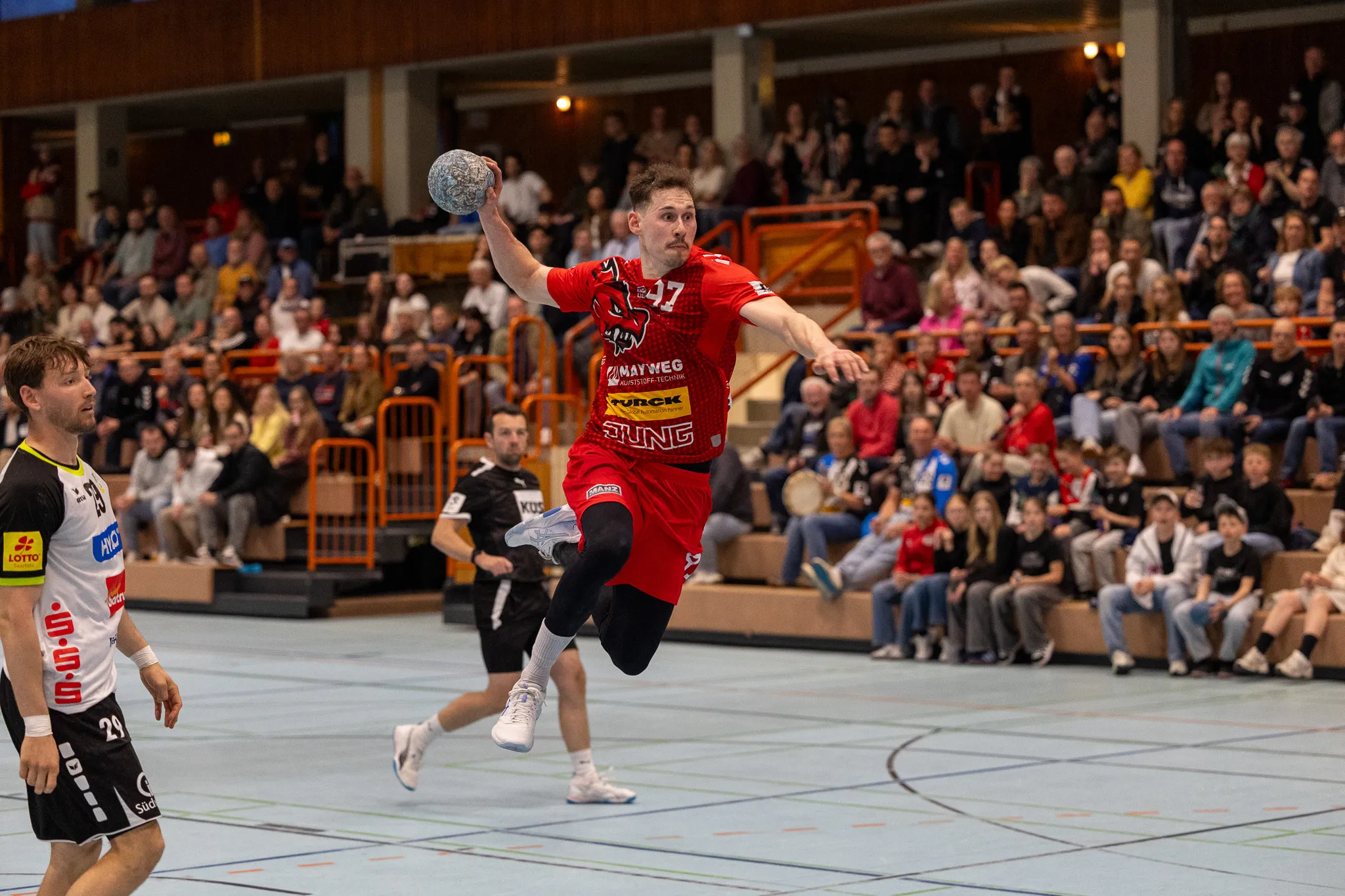Handballspieler in rotem Trikot springt zum Wurf, während ein Gegenspieler in schwarzem Trikot ihn verteidigt. Im Hintergrund eine volle Tribüne.