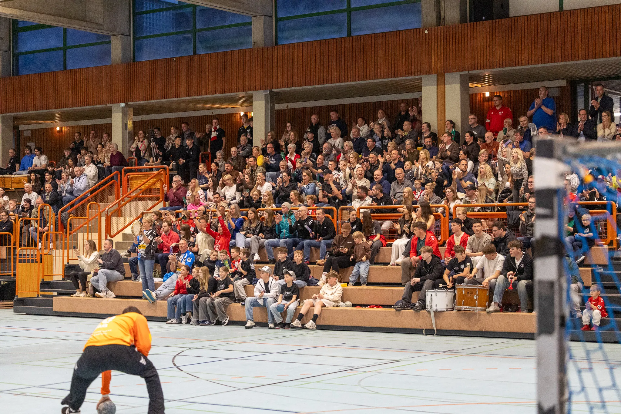 Zuschauer auf Tribünen beobachten ein Handballspiel in einer Sporthalle. Ein Spieler in orangefarbener Kleidung bereitet sich auf dem Spielfeld vor.