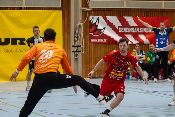 Handballspieler in roten Trikots im Angriff, während ein Torwart in Gelb und Schwarz versucht, den Schuss zu blocken. Im Hintergrund eine Werbebande und weitere Spieler.
