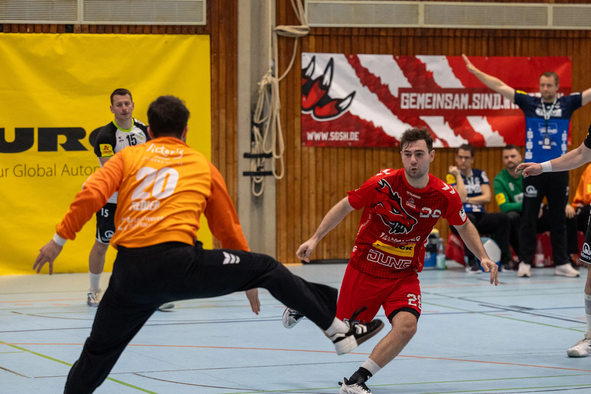 Handballspieler in roten Trikots im Angriff, während ein Torwart in Gelb und Schwarz versucht, den Schuss zu blocken. Im Hintergrund eine Werbebande und weitere Spieler.