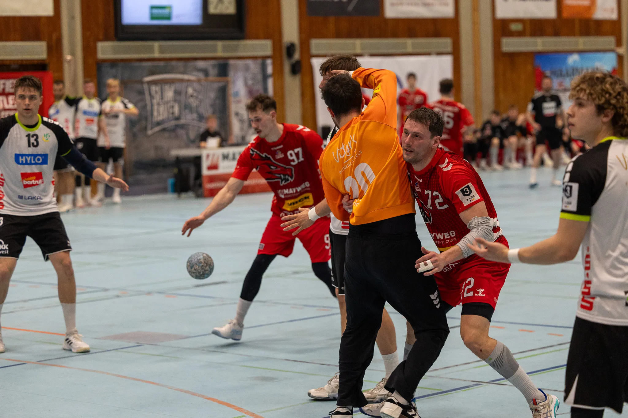 Zwei Handballspieler kämpfen um den Ball, während andere Spieler im Hintergrund stehen. Der Spieler in Schwarz hält den Ball, während sein Gegner in Rot und Gelb versucht, ihn zu blockieren.