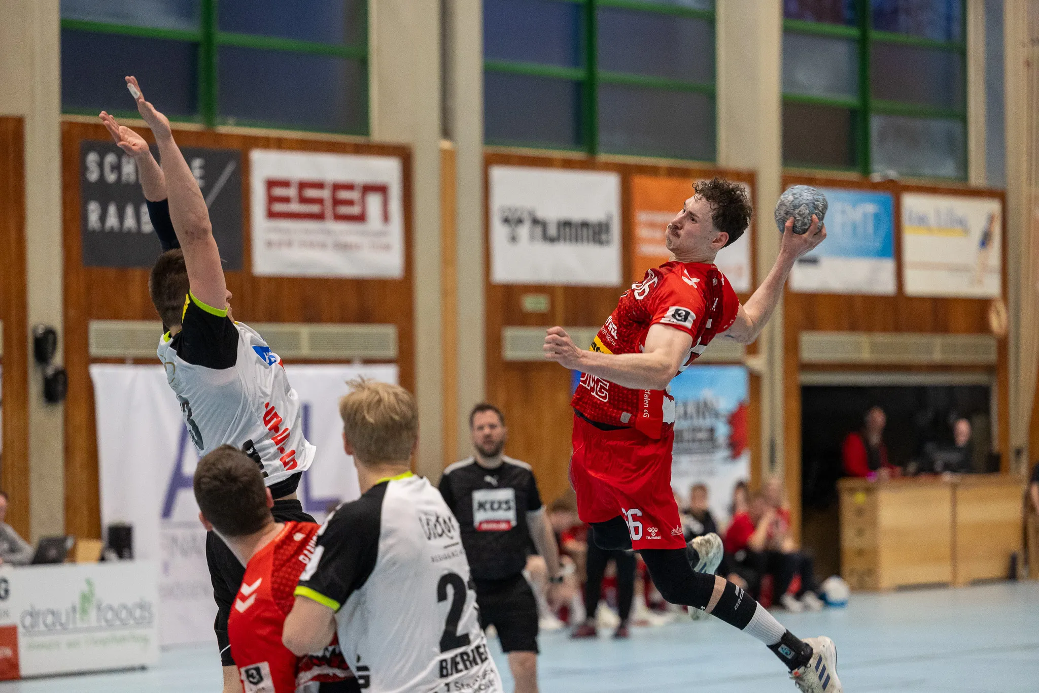 Handballspieler in roten Trikots springt zum Wurf, während ein Gegenspieler in Schwarz-Gelb versucht, ihn zu blocken. Im Hintergrund weitere Spieler und Zuschauer in einer Sporthalle.