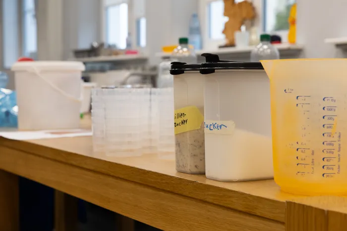 Kunststoffbehälter mit Etiketten und ein gelber Messbecher auf einem Holztisch.