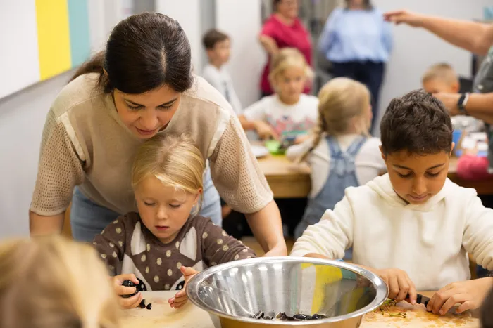 Eine Betreuerin unterstützt ein Kind bei einer Tätigkeit an einem Tisch mit einer Metallschüssel, während im Hintergrund weitere Kinder in einem Gruppenraum sind.