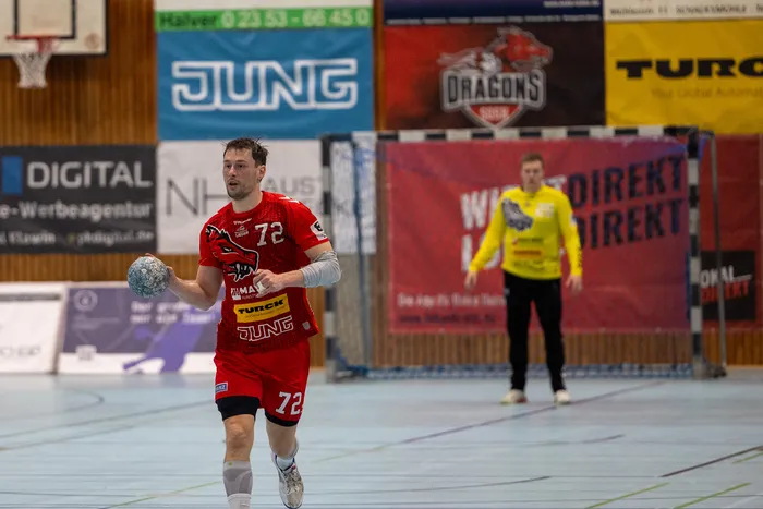 Handballspieler in rotem Trikot mit der Nummer 72 wirft den Ball während eines Spiels. Im Hintergrund ein Tor und eine weitere Spielerin in gelbem Trikot.