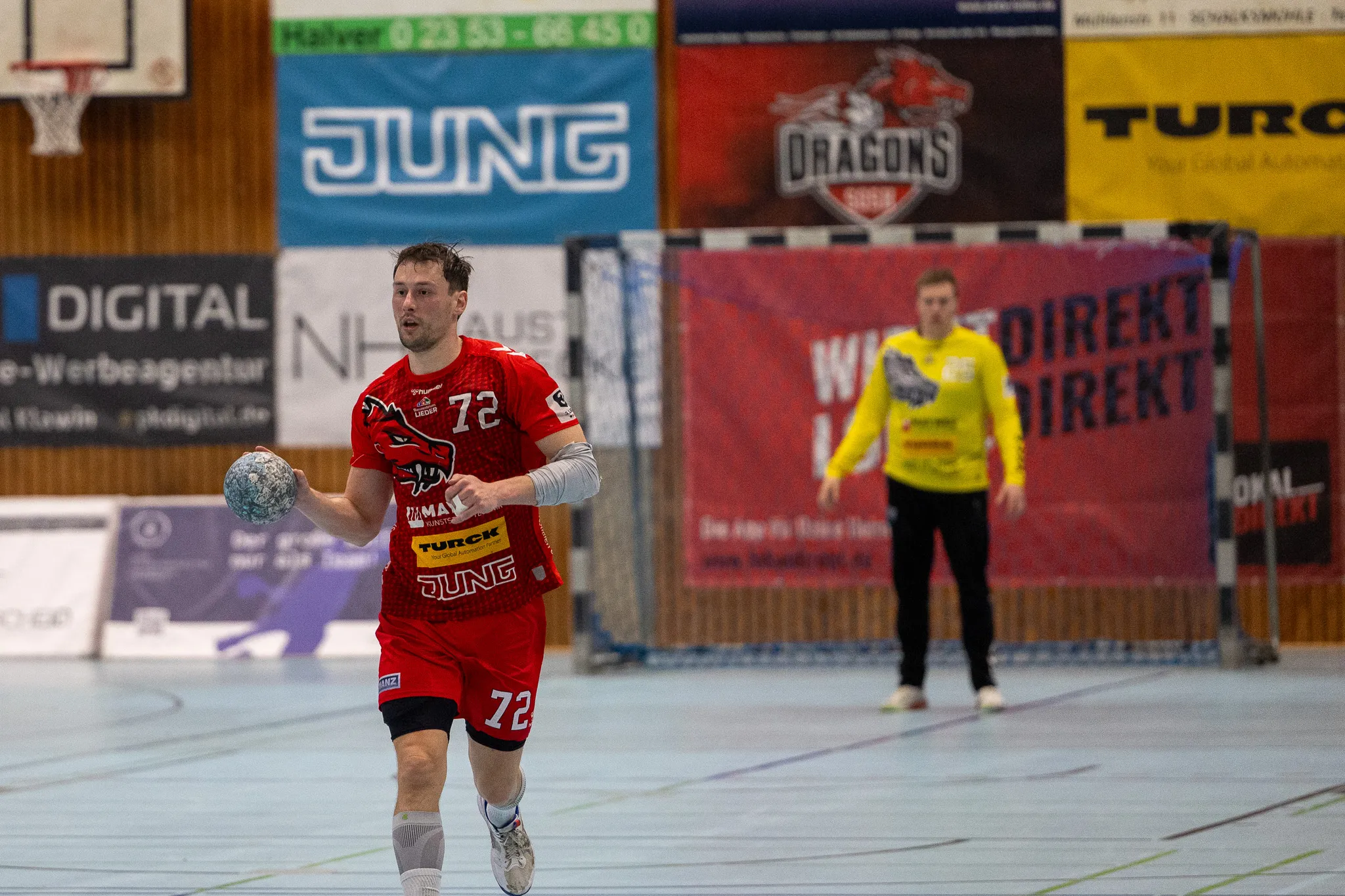Handballspieler in rotem Trikot mit der Nummer 72 wirft den Ball während eines Spiels. Im Hintergrund ein Tor und eine weitere Spielerin in gelbem Trikot.