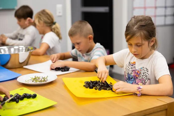 Ein Mädchen schneidet Weintrauben auf einem gelben Schneidebrett, während im Hintergrund weitere Kinder am Tisch sitzen und ebenfalls Obst schneiden.